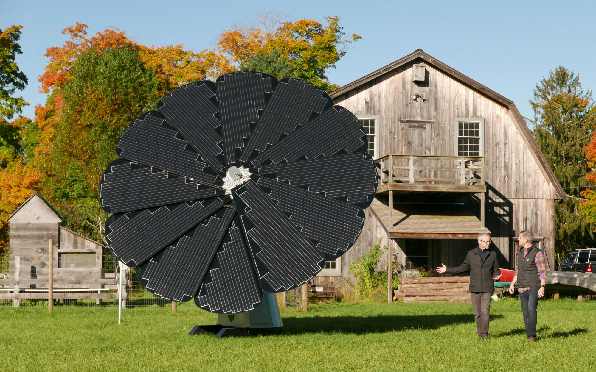 Two Men Stand Outside a Wooden Barn With a SmartFlower Solar Panel Out Front